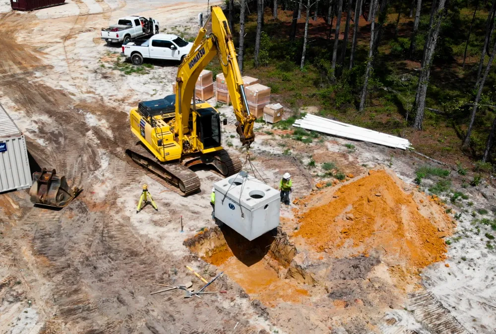 Excavator and crew setting a concrete septic tank on a wooded residential lot