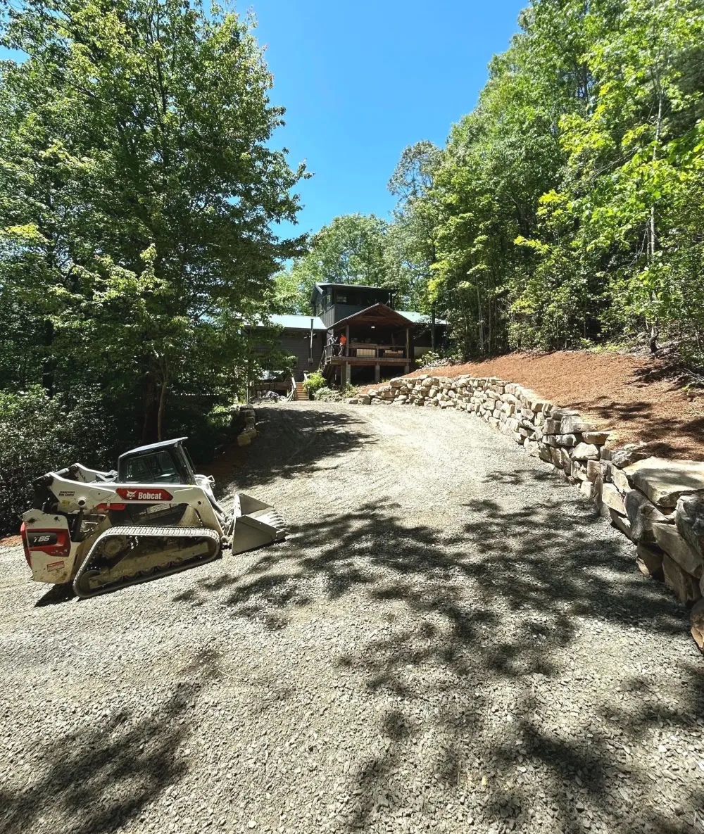Compact track loader spreading fresh gravel on a new driveway beside a stacked stone retaining wall