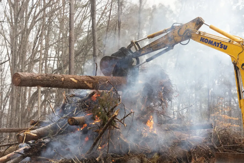 Komatsu excavator lifting a felled log onto a smoking burn pile in the woods
