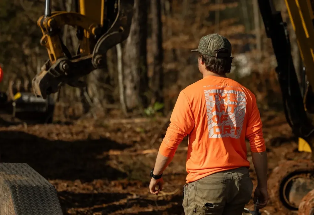 Crew member in an orange Art of Dirt shirt guiding an excavator working in the treeline